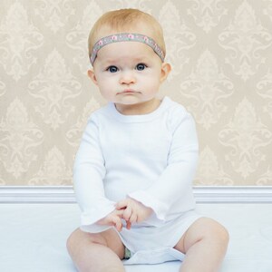 May include: A baby wearing a white long-sleeved shirt with a floral headband. The baby is sitting on a white surface with a patterned background.