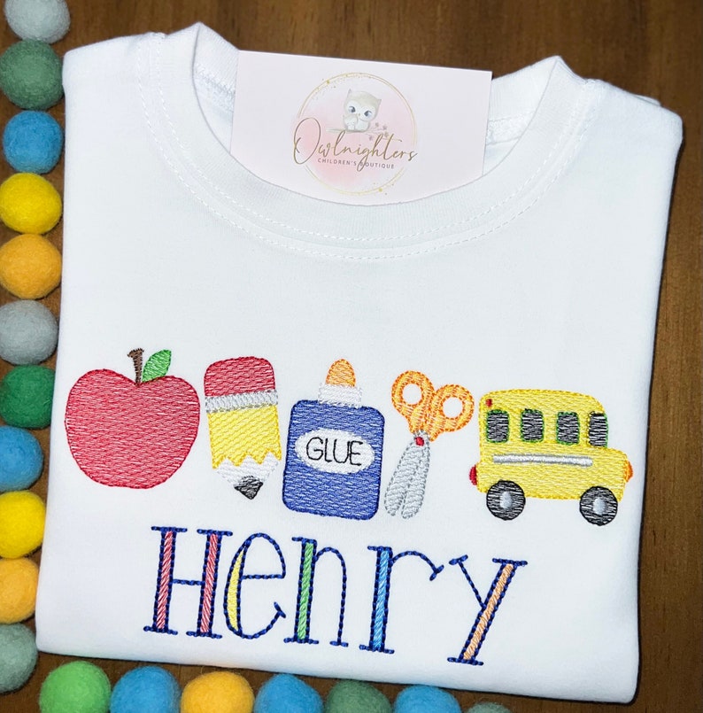May include: White t-shirt with a colourful embroidered design of a red apple, yellow pencil, blue glue bottle, orange scissors, and a yellow school bus. The name "Henry" is embroidered below the design in rainbow colours.