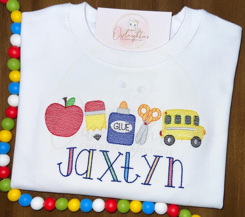 May include: White t-shirt with a red apple, yellow pencil, blue glue bottle, scissors, and a yellow school bus embroidered on it. The name "Jaxtyn" is embroidered below the school supplies.