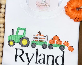 May include: White t-shirt with an embroidered green tractor pulling a wagon filled with orange pumpkins. The name "Ryland" is embroidered in black script below the tractor. A small tag with the text "Owlknighters Children's Boutique" is visible.