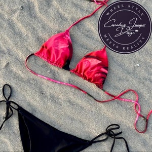 May include: A red floral print bikini top with black straps and a black bikini bottom with adjustable ties. The bikini is on a sandy beach. The image also includes a circular logo with the text "Where Style Meets Reality" and "Cascading Juniper Design TM".