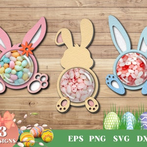 Puede incluir: Tres soportes para dulces con forma de conejo de Pascua, uno rosa, uno azul y uno amarillo. Cada soporte tiene una ventana de plástico transparente para mostrar los dulces. Los soportes están recortados en madera y tienen un acabado brillante. La imagen también incluye un fondo de hierba verde con huevos de Pascua.