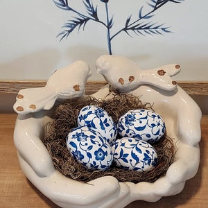 May include: A white ceramic decorative bowl shaped like hands, holding a nest of dried moss and four blue and white patterned eggs. Two white ceramic birds perch on the rim. The background features a blue and white floral design.