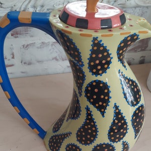 May include: A decorative ceramic pitcher with a unique, curved shape. The pitcher features a light yellow base adorned with black paisley shapes outlined in blue and filled with orange dots. The handle is blue with orange accents, and the lid has a pink and black design.