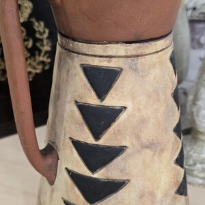 May include: A handcrafted ceramic pitcher with a brown top and handle, and a cream-colored body. The body is decorated with black triangle patterns. The rim has a scalloped design. The pitcher is approximately 8 inches tall.