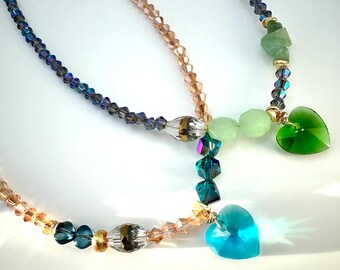 Puede incluir: Dos collares superpuestos con cuentas de cristal en tonos azules, verdes y rosas. Los collares presentan colgantes en forma de corazón en verde y azul.