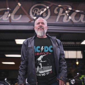 May include: A man wearing a black t-shirt with a graphic of the band AC/DC in Japanese text and a photo of the band performing on stage. The text reads "AC/DC 日本 の ツアー" and "TOKYO '81".