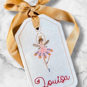 May include: A white gift tag with a gold ribbon bow. The tag features an embroidered ballerina in a pink tutu and the name "Louisa" in red script. The ballerina has a gold crown and fairy wings.