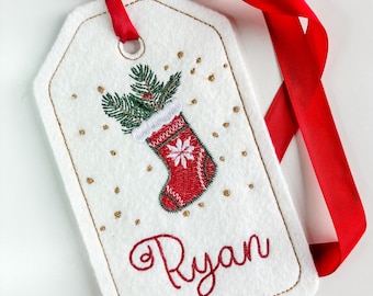 May include: White felt Christmas gift tag with gold stitching, featuring an embroidered red stocking with a green sprig of holly and gold glitter. The tag is tied with a red ribbon and has the name "Ryan" embroidered in red.