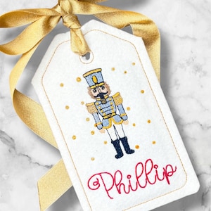 May include: A white gift tag with a gold ribbon bow. The tag features an embroidered nutcracker soldier in blue, gold, and black, with the name "Phillip" in red script. Gold dots are scattered around the nutcracker.