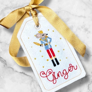 May include: White felt gift tag with a gold ribbon. The tag features an embroidered Nutcracker mouse in a blue and red uniform, holding a sword. The word "Ginger" is embroidered in red script, with gold dots.
