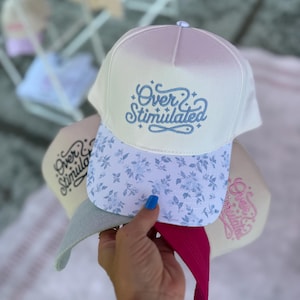 May include: A stack of baseball caps with the text "Over Stimulated" embroidered on the front. The top cap is white with a floral pattern on the brim. The brim of the cap is light blue with a floral pattern. The other caps are in shades of beige, pink, and gray.