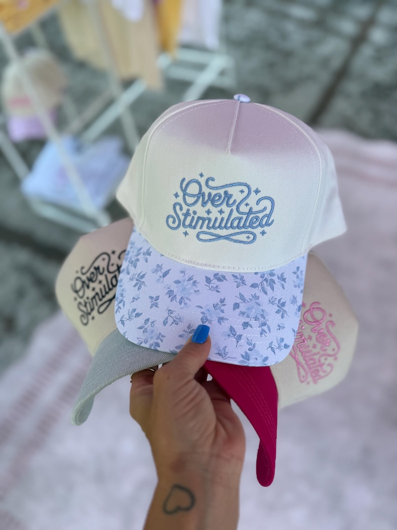 May include: A stack of baseball caps with the text "Over Stimulated" embroidered on the front. The top cap is white with a floral pattern on the brim. The brim of the cap is light blue with a floral pattern. The other caps are in shades of beige, pink, and gray.