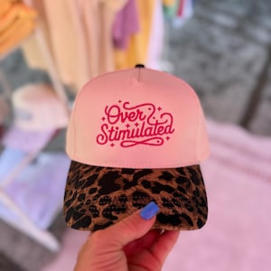 May include: A pink baseball cap with the embroidered text "Over Stimulated" in a cursive font. The brim is patterned with a brown and black leopard print. The cap is held up by a hand with blue nail polish.