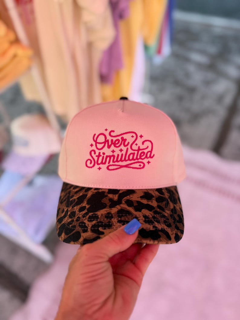 May include: A pink baseball cap with the embroidered text "Over Stimulated" in a cursive font. The brim is patterned with a brown and black leopard print. The cap is held up by a hand with blue nail polish.