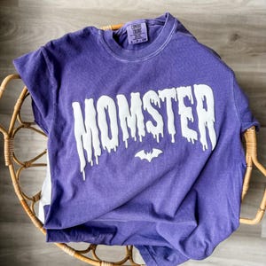 May include: A purple short-sleeved t-shirt with a crew neckline. The front features the white text MOMSTER in a dripping font style, with a small white bat icon centered below the text. The inner neck label reads Comfort Colors size S.