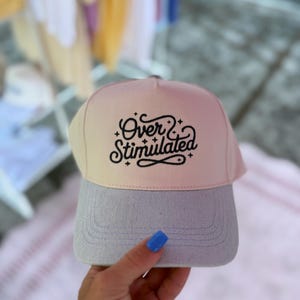 May include: A baseball cap with a light pink and gray color scheme. The cap features the text "Over & Stimulated" in a black script font. The brim is gray, and the crown transitions from light pink to beige.