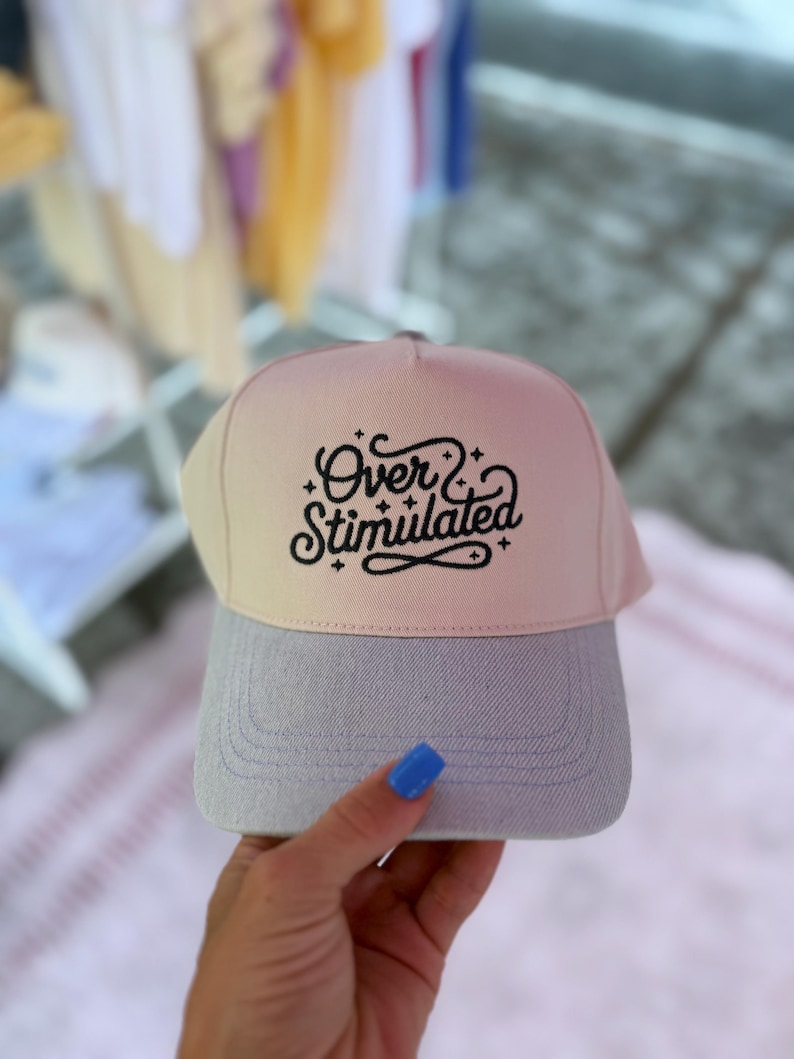 May include: A baseball cap with a light pink and gray color scheme. The cap features the text "Over & Stimulated" in a black script font. The brim is gray, and the crown transitions from light pink to beige.
