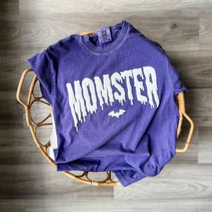 May include: A purple short-sleeve t-shirt with a crew neckline. The front features the word MOMSTER printed in a white, dripping font style, with a small white bat icon centered below the text. A Comfort Colors brand label is visible at the neck.