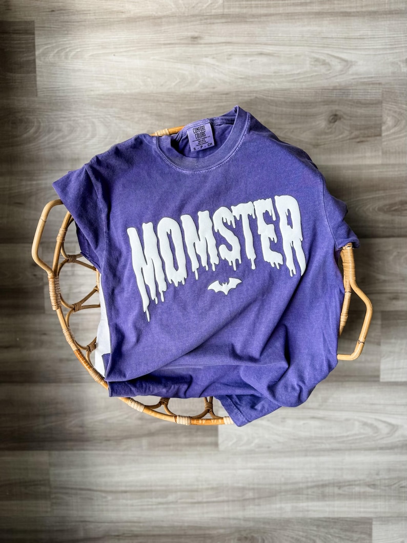 May include: A purple short-sleeve t-shirt with a crew neckline. The front features the word MOMSTER printed in a white, dripping font style, with a small white bat icon centered below the text. A Comfort Colors brand label is visible at the neck.