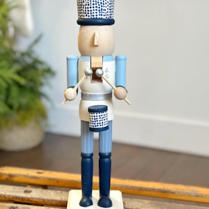 May include: A wooden nutcracker soldier in a drum major uniform. The figure has a light wood head and a blue and white uniform. The hat and drum are navy blue with white polka dots. The figure stands on a white base.