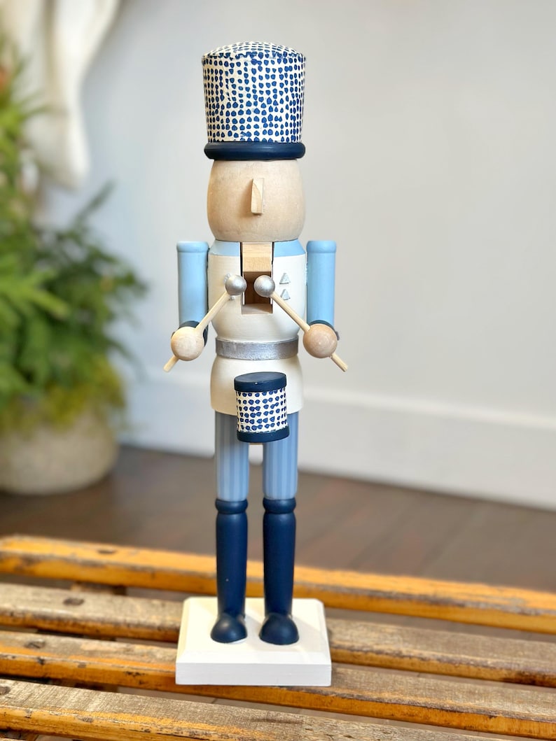 May include: A wooden nutcracker soldier in a drum major uniform. The figure has a light wood head and a blue and white uniform. The hat and drum are navy blue with white polka dots. The figure stands on a white base.
