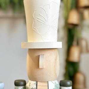 May include: A wooden nutcracker figurine with a white top hat featuring a floral design. The figurine has a light wood face with a small rectangular ear. The body is decorated with a green floral pattern.