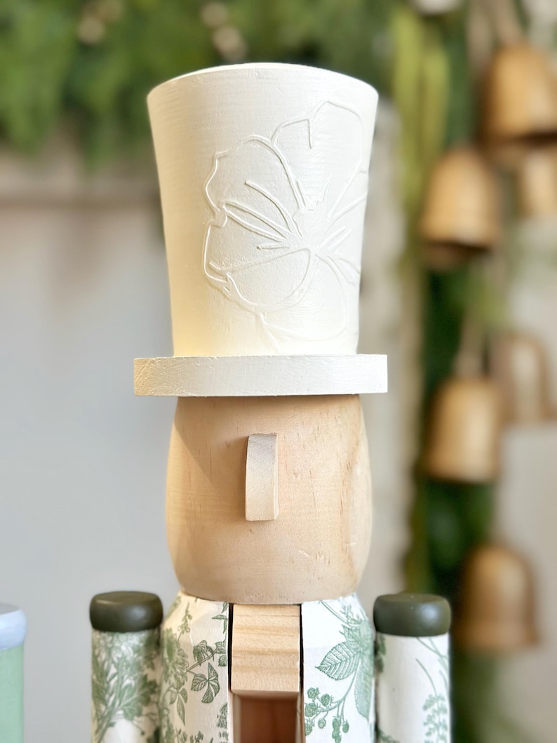May include: A wooden nutcracker figurine with a white top hat featuring a floral design. The figurine has a light wood face with a small rectangular ear. The body is decorated with a green floral pattern.