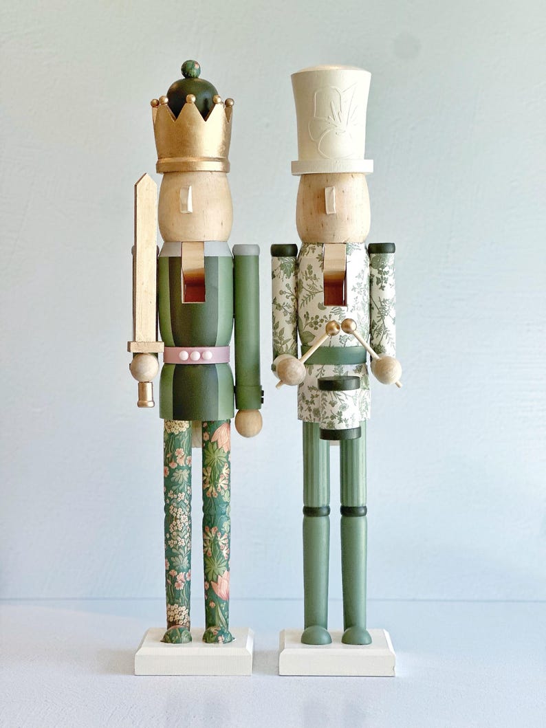 May include: Two decorative wooden nutcrackers. One wears a gold crown and holds a sword, the other a white top hat and drumsticks. Both have green and floral patterned details, standing on white bases.