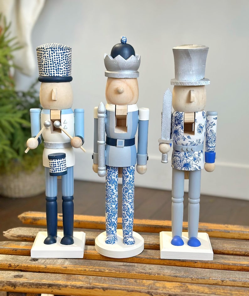 May include: Three wooden nutcrackers in a row, each with a unique design. The nutcrackers are painted in shades of blue, white, and silver. One holds drumsticks, another a staff, and the third a sword. They stand on white bases.