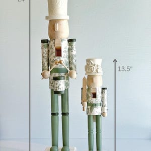 May include: Two decorative wooden nutcrackers. The larger one is 61 cm tall, and the smaller one is 34 cm tall. Both have a light wood head and a white hat. The bodies are decorated with a green and white floral pattern. The legs are green.