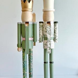 May include: Two decorative wooden nutcrackers. One wears a gold crown and green uniform with floral patterned legs. The other wears a white top hat and a white and green floral patterned uniform. Both stand on white bases.