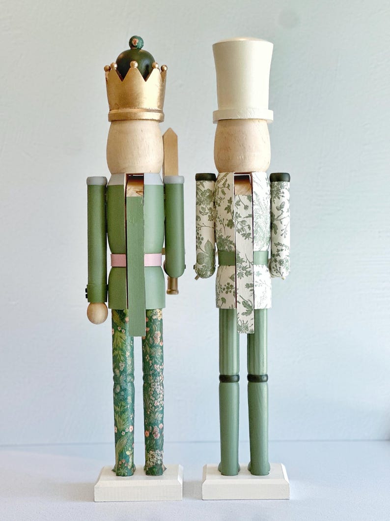 May include: Two decorative wooden nutcrackers. One wears a gold crown and green uniform with floral patterned legs. The other wears a white top hat and a white and green floral patterned uniform. Both stand on white bases.