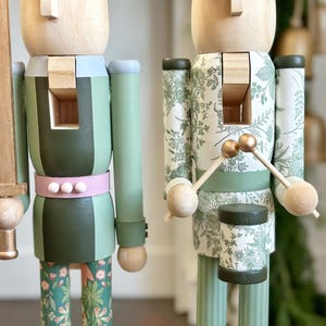 May include: Two wooden nutcrackers. One holds a gold sword, and the other holds drumsticks over a drum. The nutcrackers are painted in shades of green, with floral and striped patterns. One has a pink belt with white dots.