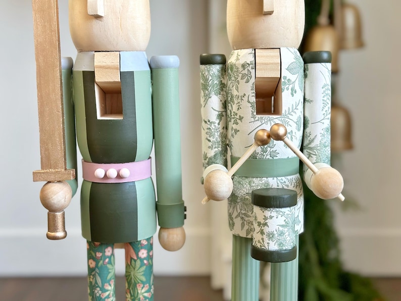 May include: Two wooden nutcrackers. One holds a gold sword, and the other holds drumsticks over a drum. The nutcrackers are painted in shades of green, with floral and striped patterns. One has a pink belt with white dots.