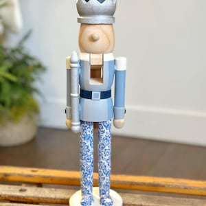 May include: A wooden nutcracker soldier with a silver crown and a dark blue hat. The soldier wears a light blue uniform with a dark blue belt and white and blue patterned pants. The nutcracker stands on a white circular base.