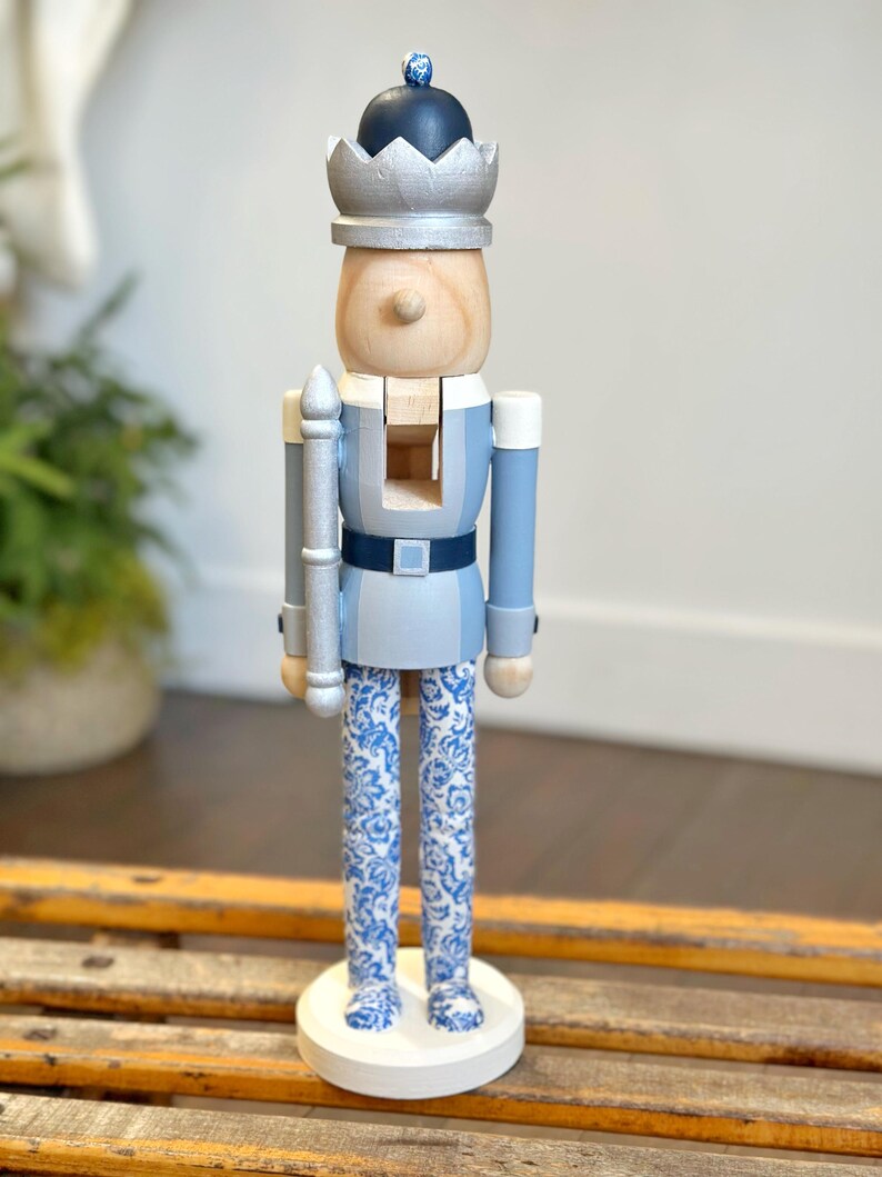 May include: A wooden nutcracker soldier with a silver crown and a dark blue hat. The soldier wears a light blue uniform with a dark blue belt and white and blue patterned pants. The nutcracker stands on a white circular base.
