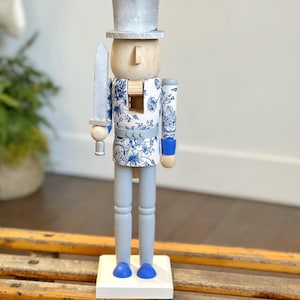 May include: A decorative wooden nutcracker figurine. The figurine has a silver top hat and sword, a light wood face, and blue and white floral patterned clothing. The legs and base are painted light gray and white, respectively. The nutcracker stands on a white base.