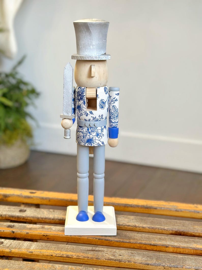 May include: A decorative wooden nutcracker figurine. The figurine has a silver top hat and sword, a light wood face, and blue and white floral patterned clothing. The legs and base are painted light gray and white, respectively. The nutcracker stands on a white base.