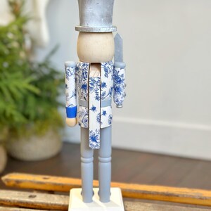 May include: A decorative wooden nutcracker figurine. The figurine has a silver top hat, a light wood head, and a blue and white floral patterned jacket and scarf. The legs and base are painted gray and white, respectively. A silver knife is held at the side.
