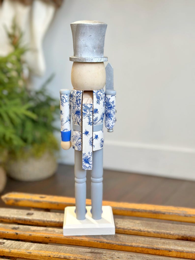 May include: A decorative wooden nutcracker figurine. The figurine has a silver top hat, a light wood head, and a blue and white floral patterned jacket and scarf. The legs and base are painted gray and white, respectively. A silver knife is held at the side.