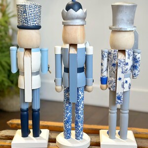 May include: Three decorative wooden nutcrackers in shades of blue, white, and gray. Each nutcracker has a unique design, including patterned hats and clothing. They stand on white bases, adding a touch of festive charm.