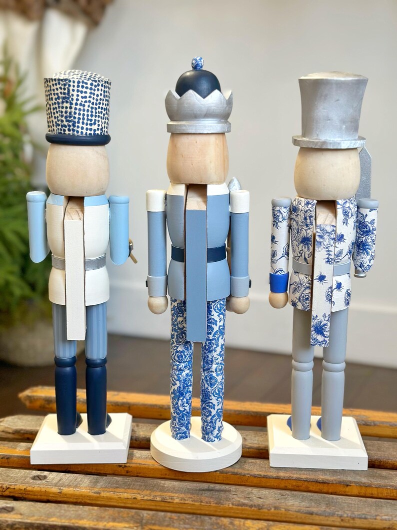 May include: Three decorative wooden nutcrackers in shades of blue, white, and gray. Each nutcracker has a unique design, including patterned hats and clothing. They stand on white bases, adding a touch of festive charm.