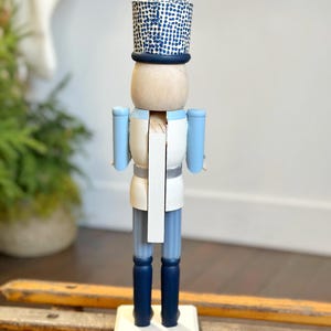 May include: A wooden nutcracker figurine in a soldier's uniform. The figurine has a light blue jacket, dark blue pants and boots, and a white shirt. The hat is blue with a white dotted pattern. The nutcracker stands on a white base.