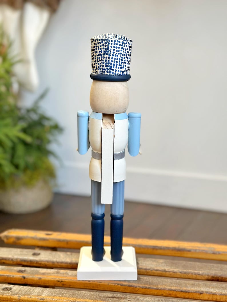 May include: A wooden nutcracker figurine in a soldier's uniform. The figurine has a light blue jacket, dark blue pants and boots, and a white shirt. The hat is blue with a white dotted pattern. The nutcracker stands on a white base.