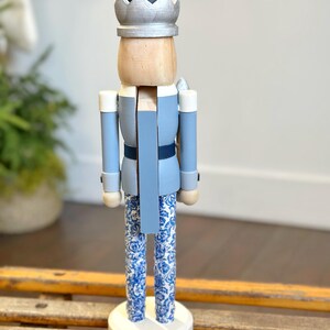 May include: A decorative wooden nutcracker figurine. The figurine is painted in shades of blue, with a silver crown and a black hat. The legs feature a blue and white floral pattern. The nutcracker stands on a white circular base.