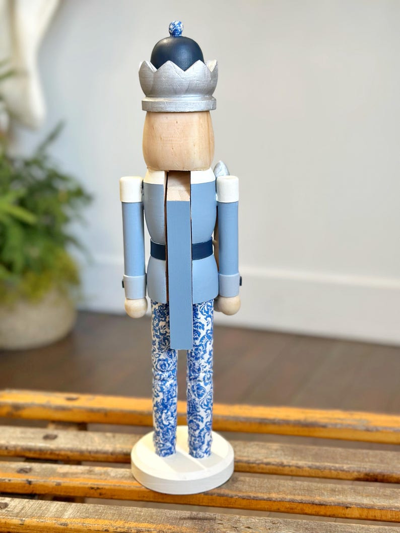 May include: A decorative wooden nutcracker figurine. The figurine is painted in shades of blue, with a silver crown and a black hat. The legs feature a blue and white floral pattern. The nutcracker stands on a white circular base.