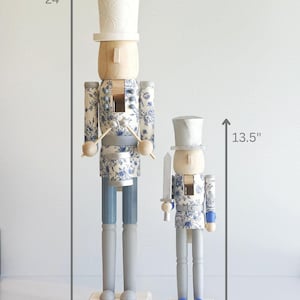 May include: Two wooden nutcrackers, one 24 inches tall and the other 13.5 inches tall. Both have a light wood head and a white hat. The bodies are decorated with a blue floral pattern. One holds drumsticks, the other a sword.