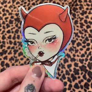 Puede incluir: Una pegatina holográfica de un diablo de dibujos animados con cuernos rojos y un atuendo rojo con capucha. El diablo sostiene una piruleta en la boca y lleva una pajarita roja.