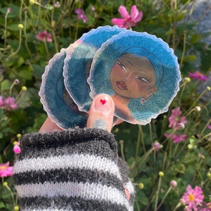 May include: A stack of round stickers featuring a cartoon illustration of a person with blue hair and a light skin tone. The stickers are held in front of a background of pink flowers and green foliage.